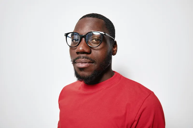 Portrait of African American man with dark skin wearing glasses and red sweater looking at camera with confident expression on white background. Studio shot with casual style.