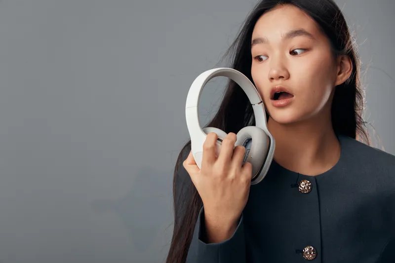 Young woman with long dark hair holding white headphones looking surprised or curious against grey background. Studio portrait of female expressing emotion with technology device in hand.