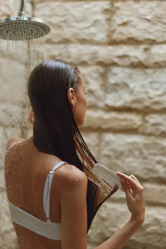 Woman combing wet hair under warm outdoor rain shower, natural skin, stone wall background, peaceful moment, summer relaxation.