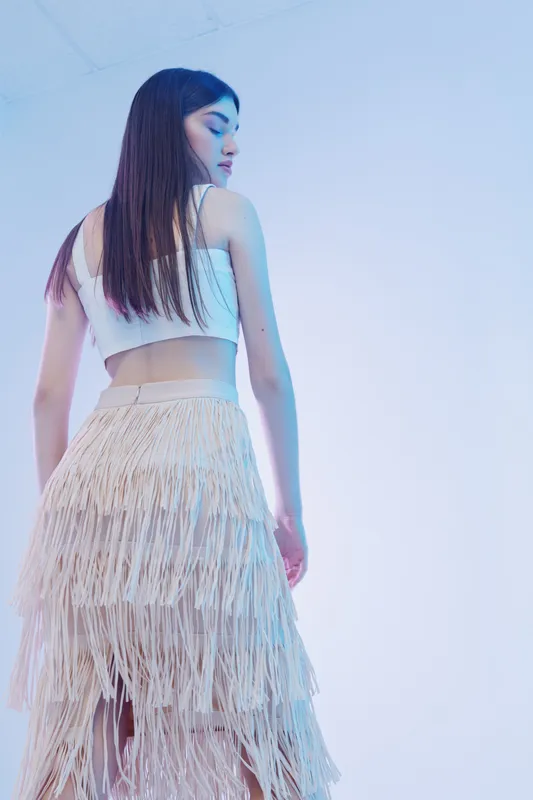 Young woman in a white crop top and fringed skirt poses sideways in a studio with soft blue lighting. Concept of woman beauty fashion and modern style.