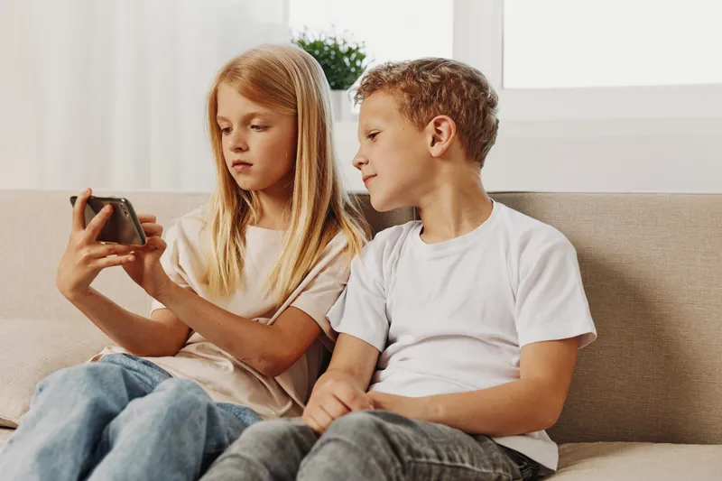 Two children sitting on a couch indoors focused on a smartphone screen together. Casual clothing with natural light from window in the background.