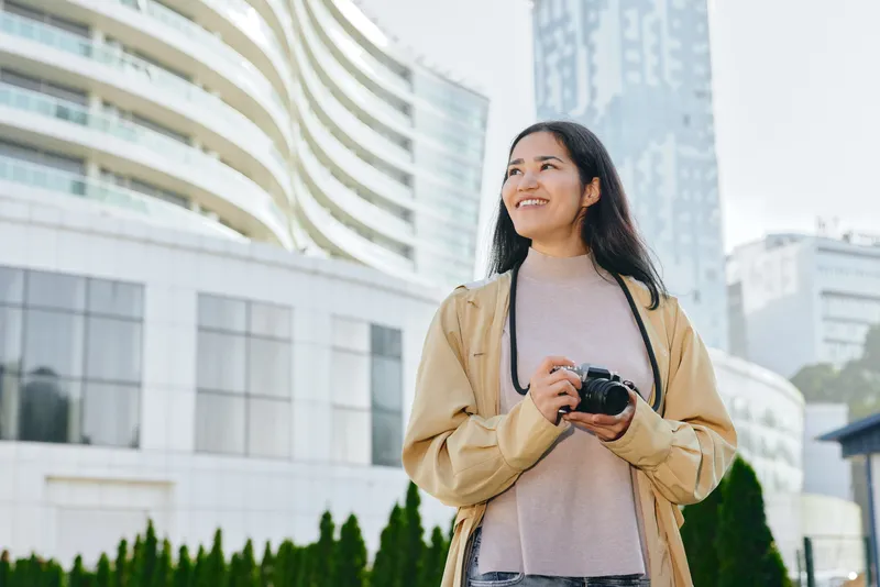 Camera, photography, outdoor, urban, casual, chic, scene of a smiling female shutterbug holding a camera in hand amid modern buildings and greenery, conveying creativity, mobility, and city lifestyle