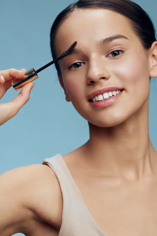 Young woman applying mascara with a cheerful expression against a serene blue background, showcasing beauty and self care, ideal for cosmetics marketing