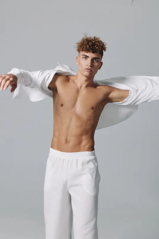 Young man with curly hair wearing white pants and open white shirt posing against gray background. Male model showing toned body and confident expression in studio.