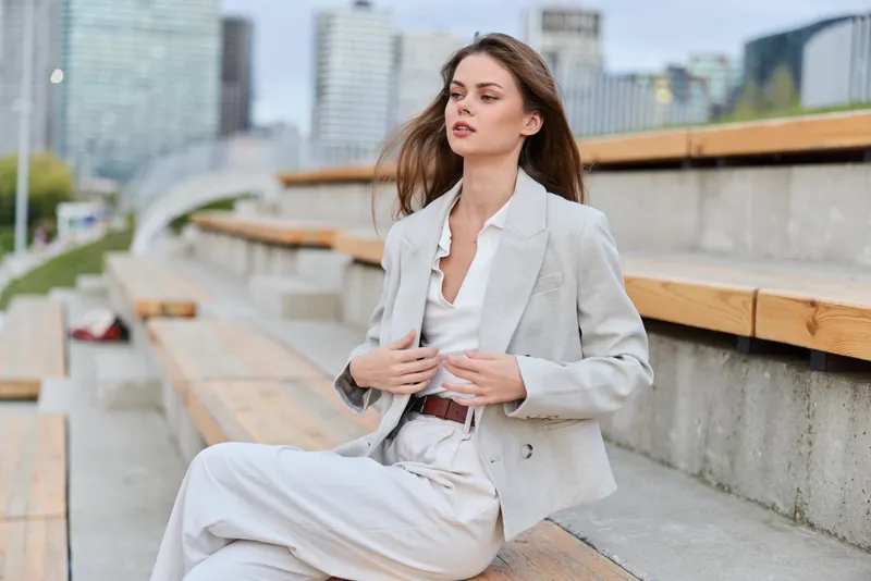 Young woman in light beige business suit sitting outdoors on wooden benches with cityscape background, confident pose, natural makeup, spring season
