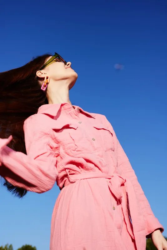Fashionable young woman in pink dress against bright blue sky, expressing joy and confidence while showcasing a vibrant summer style