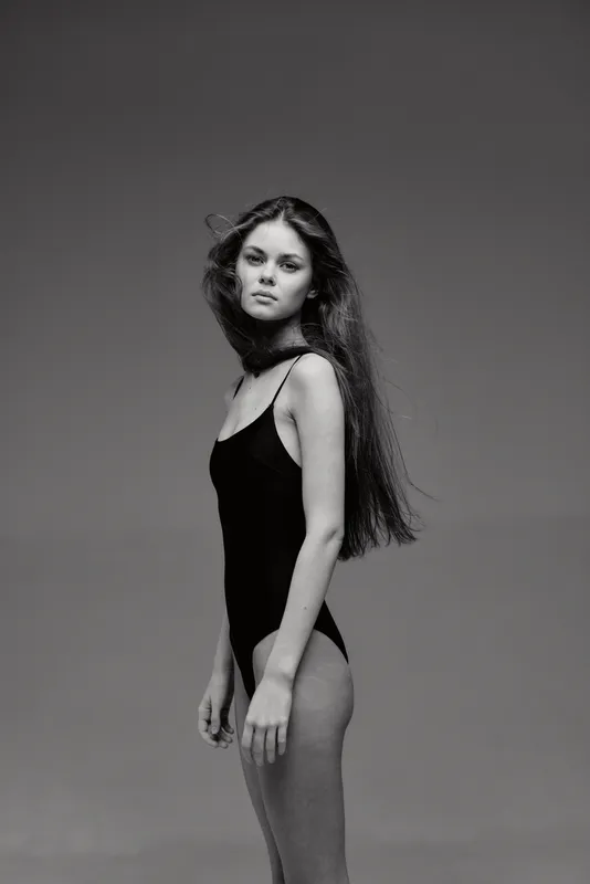 Black and white portrait of a young woman in a black swimsuit with long hair and a serious expression.