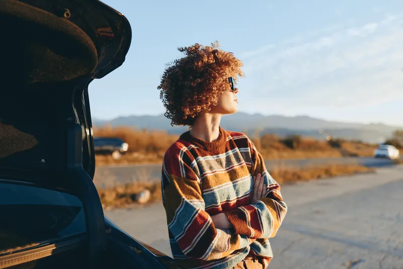 Woman lifestyle portrait beside a car on an open road, curly hair in the wind, warm sunlight and distant mountains behind. Relaxed travel moment outdoors.