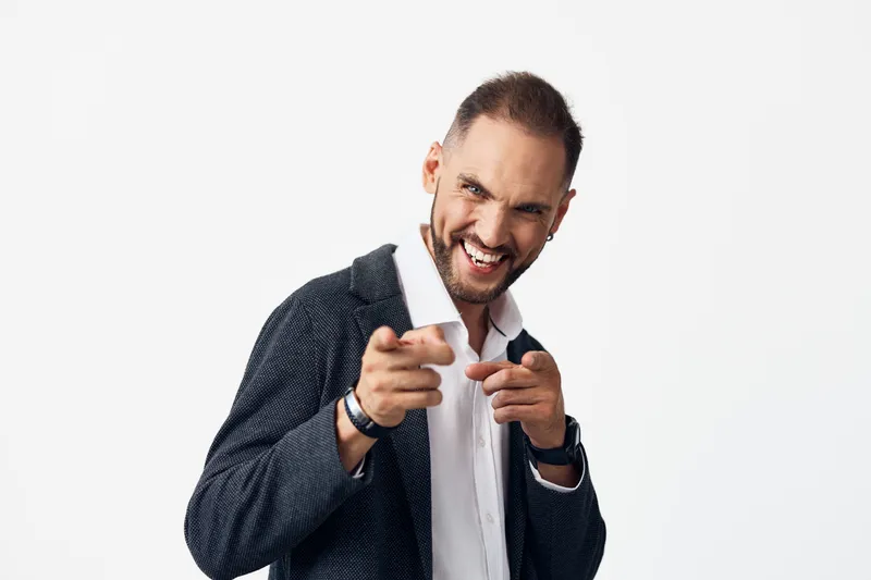 A confident business professional man in a dark blazer smiles and points toward the camera against a clean colored isolated backdrop.