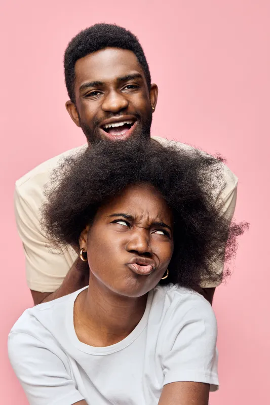 Joyful couple laughing together with playful expressions against a pastel pink background, showcasing youthfulness and spontaneity