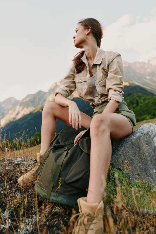A young woman in casual outdoor clothing sits on a rock with a backpack, looking to the side in a mountainous landscape during daytime. Lifestyle and adventure concept.
