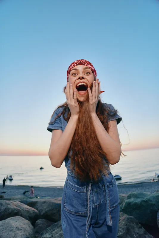 Lifestyle woman in modern boho-western denim street style stands on rocky beach with film color effect, expressing joyful surprise at sunset sky and calm sea background.