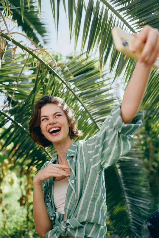 Joyful woman taking a selfie under lush palm leaves, smiling broadly as she captures a bright outdoor moment with sunny vibes.