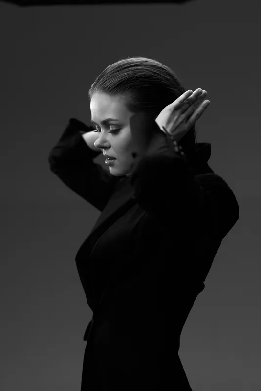 Black and white portrait of a focused woman adjusting hair in stylish dark clothing with dramatic lighting and minimalist background.