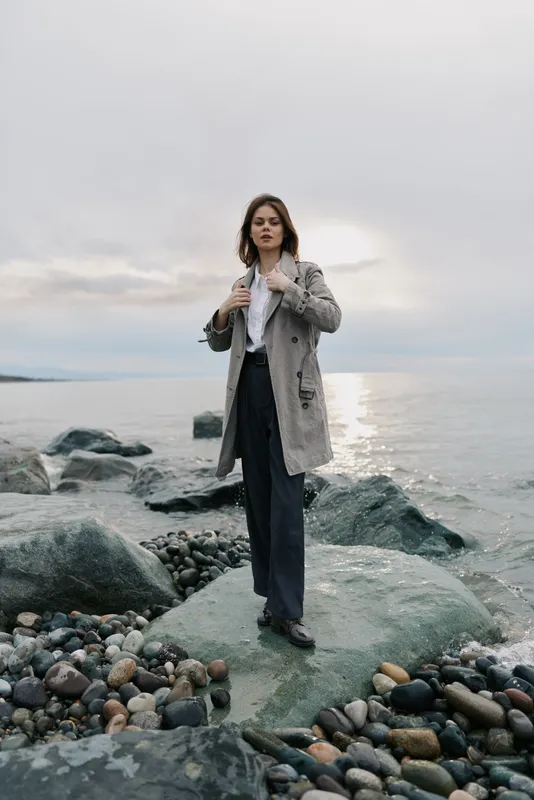 Young woman in a long coat stands on rocky shore by the sea during cloudy weather. Natural light highlights the calm water and scattered stones. Concept of outdoor fashion and nature.