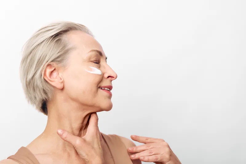Mature woman with short grey hair applies white face cream on cheek, smiling gently. Skin care and aging beauty concept with natural expression against plain white background in studio.