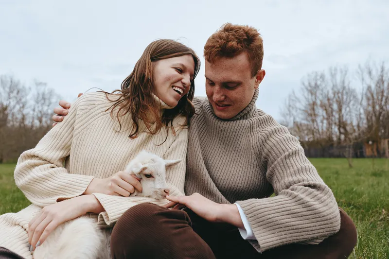 Young couple in cozy sweaters enjoys a tender moment outdoors while holding a small white baby goat on a cloudy day in a natural setting with bare trees.