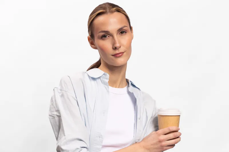 Confident young woman with natural makeup holds disposable coffee cup, wearing casual shirt and white top against plain white background. Concept of lifestyle, relaxation, and minimalism.