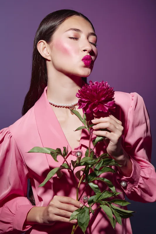 Young woman with sleek dark hair and glossy lips in a vibrant pink blouse holds a textured flower. Studio shot on violet background. Creative beauty and fashion concept.