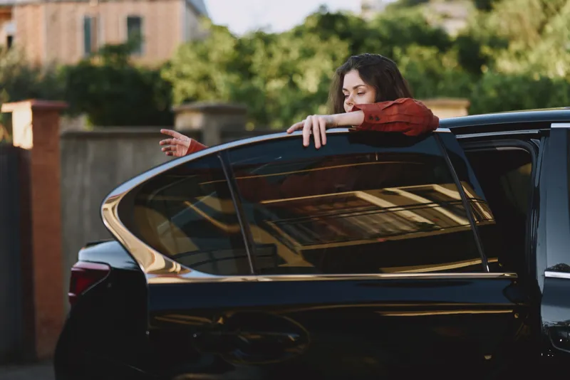 Young woman leans out of a dark car window, resting her arms on the frame with a calm, curious expression in a sunlit urban street, capturing a relaxed everyday moment