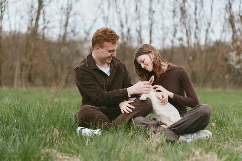 A smiling couple sits on the grass in a park, cradling a young goat kid between them. A candid lifestyle moment highlighting connection, nature, and playful companionship.