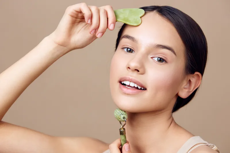 Young woman using jade roller for skin care, demonstrating self care and wellness techniques in a minimalistic beige background