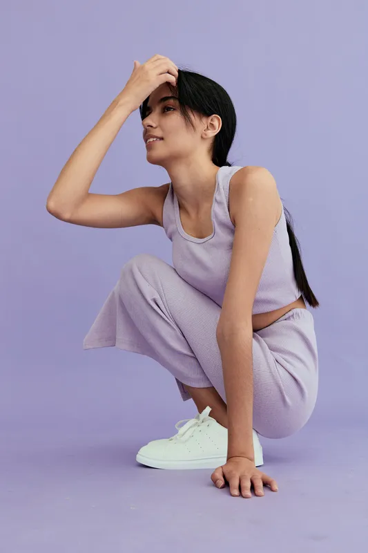 Young woman crouching in lilac activewear with white sneakers against purple background, showing relaxed pose and casual style, ideal for fitness or lifestyle themes.