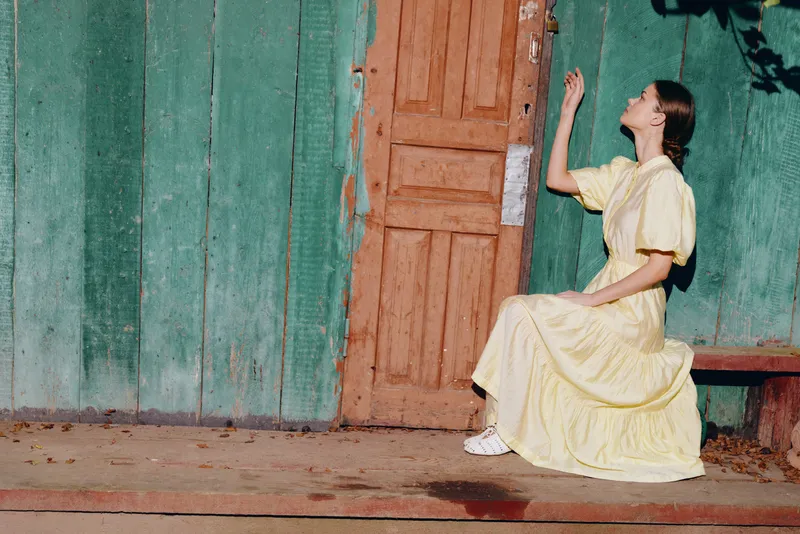 A woman in a bright yellow dress sits on a bench outdoors beside a weathered green wall and rustic door, creating a vintage outdoor scene with calm mood