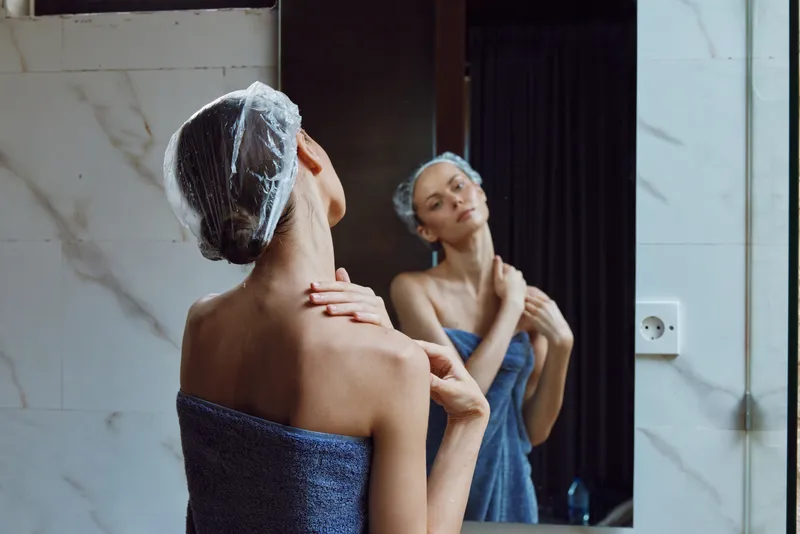 Woman in blue towel with hair cap looks at her reflection in bathroom mirror, touching shoulder in soft natural light, relaxing self-care moment indoors.