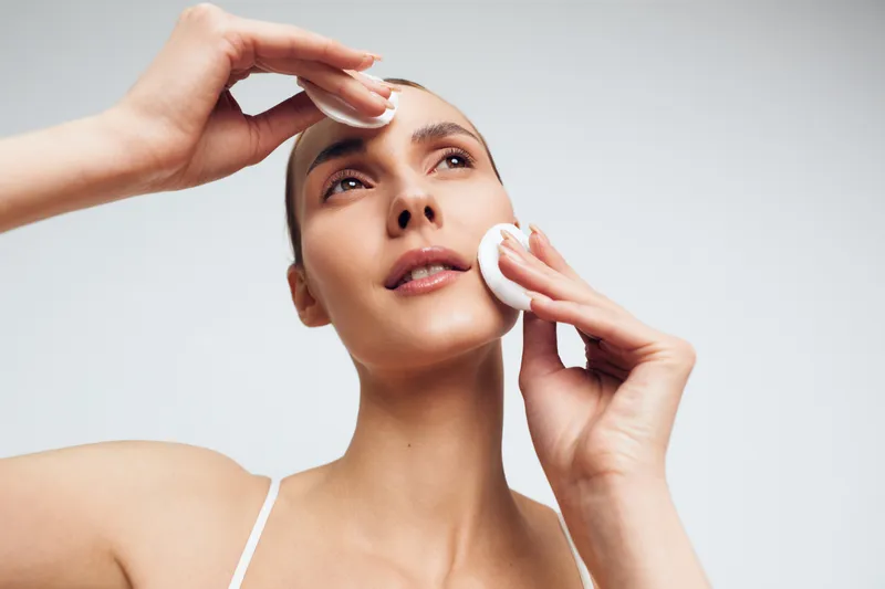 Clear portrait of a woman performing a facial cleansing routine, removing makeup with cotton pads, close up on the face, soft lighting, calm skincare moment, studio shot.