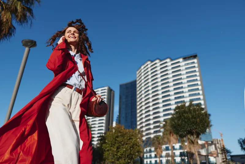 Woman outdoors in a red coat smiling in the sun, wind in her hair, urban cityscape behind, relaxed fashion pose with bright colors and clear sky, joyful energy