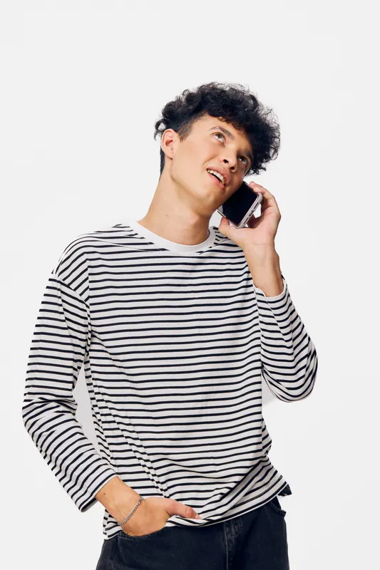 Young man talking on smartphone wearing a striped long sleeve shirt, casual clothing, curly dark hair, standing against white background, communication concept.