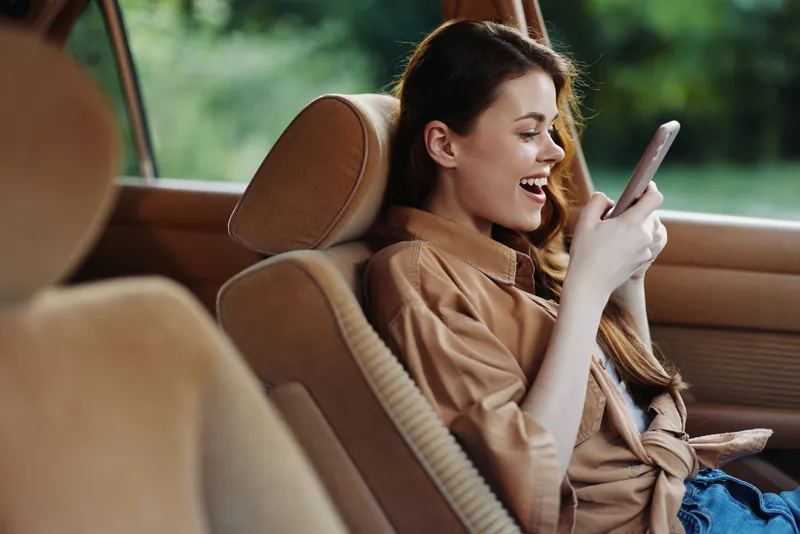 Happy young woman enjoying a moment in her car while using her smartphone, dressed in casual beige attire, surrounded by nature, embodying a carefree spirit.