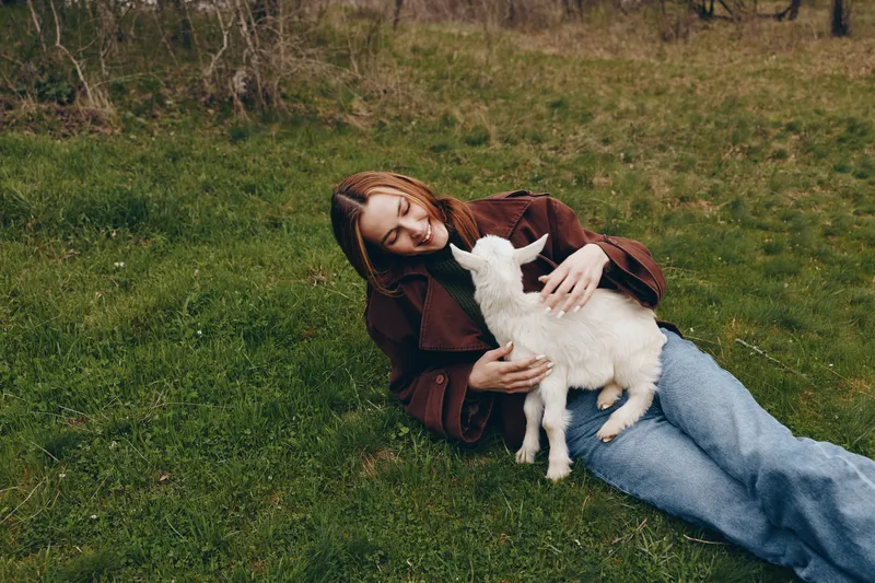 Smiling woman resting on grass, cuddling a young goat kid in a field. Outdoor lifestyle moment showing bond with animals, nature, and wellness. Soft daylight creates a warm, peaceful mood.