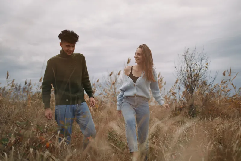 A candid outdoor portrait of a young couple walking hand in hand through tall grass, their genuine smiles and relaxed body language conveying authenticity and a natural connection.