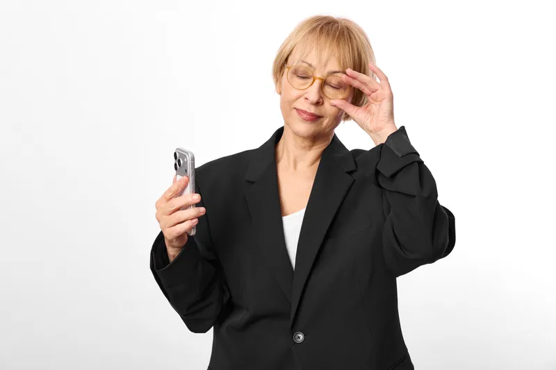 Senior woman in blazer holds smartphone, touching glasses, stressed anxious expression, worried hand gesture, isolated on white background, concept for communication stress, mental health burnout