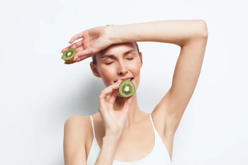Young woman with closed eyes holding two halves of fresh kiwi fruit near face, natural skincare concept, white background, healthy lifestyle, fresh and vibrant mood.