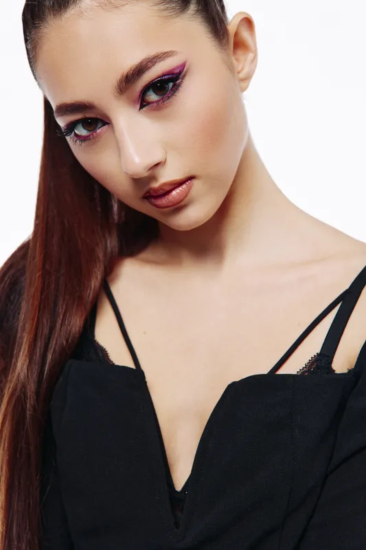 Young woman with long hair poses in a stylish black shoulder dress. Makeup features violet winged eyeliner and gloss lips. Studio shot on white background. Concept of beauty fashion creative.