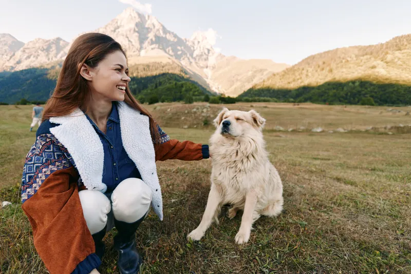 Woman dog mountain meadow outdoors pet companionship as she smiles while crouching beside fluffy dog during a hike in nature, relaxed portrait in grassy landscape and sunny daylight.