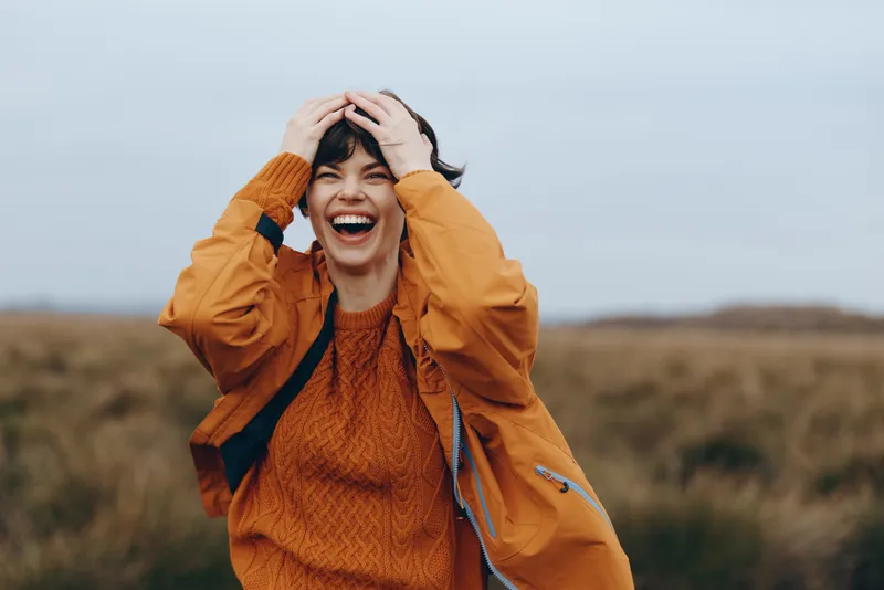 Happy woman laughing outdoors in an autumn field, hands on head, wearing orange jacket and sweater, warm nature light, lifestyle moment, calm open landscape