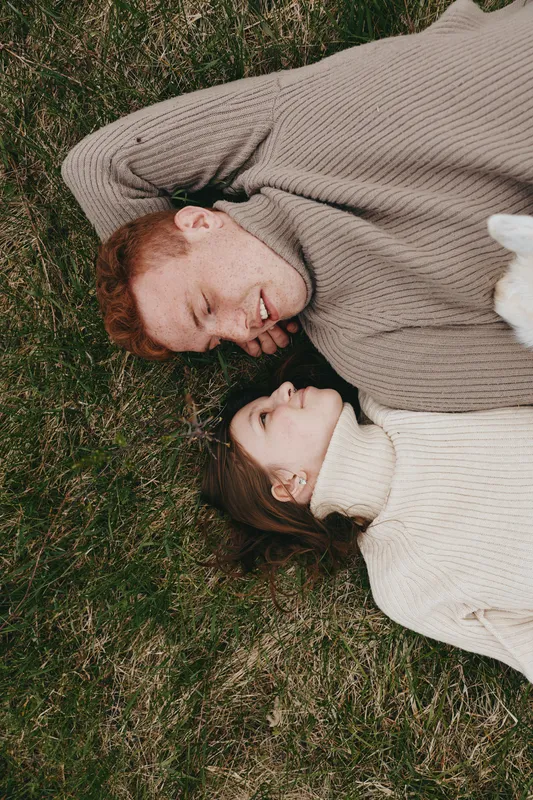 Smiling couple lying on grass in matching beige sweaters, intimate moment in natural light. Cozy outdoor lifestyle scene highlighting connection, warmth, and everyday affection.