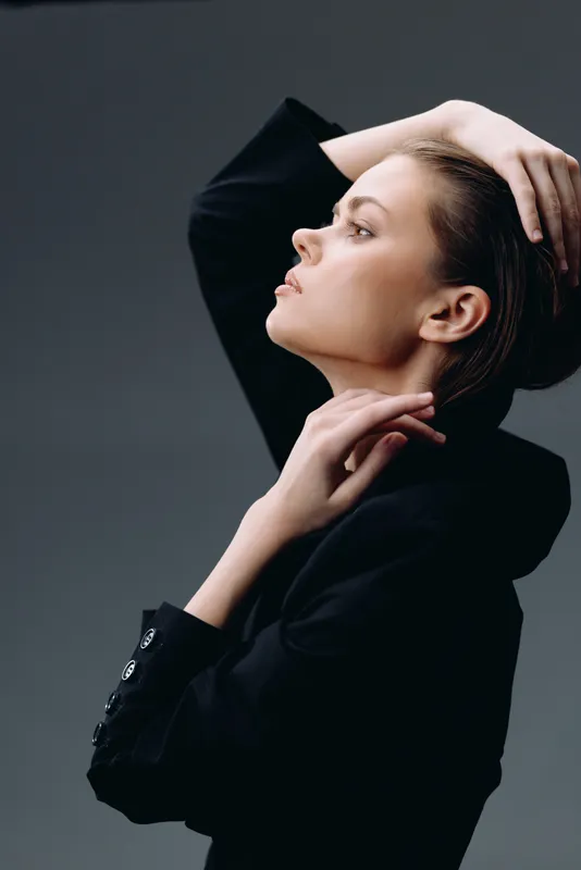 Elegant woman in black jacket posing with hands on head, showcasing sophistication and style against dark background