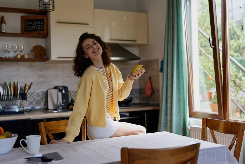 Cheerful woman indoors, lounging at a sunlit kitchen table, wearing a yellow cardigan, enjoying a fresh moment, bright natural colors, cozy home atmosphere and light breeze of everyday life captured