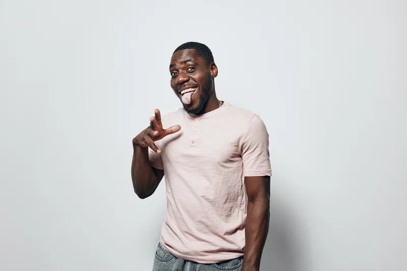 African American man with dark skin wearing light pink t shirt making playful gesture and sticking out tongue on white background. Casual, happy, young adult male portrait.
