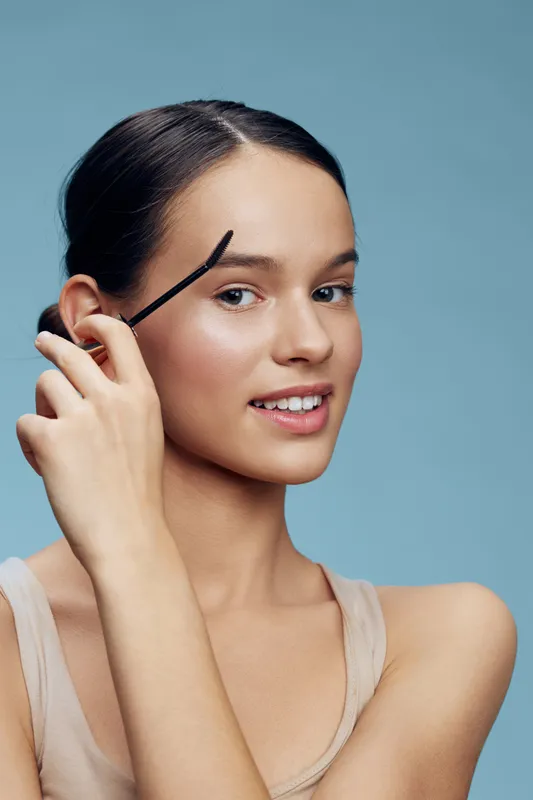 Young woman grooming her eyebrows with an eyebrow brush, showcasing a natural look and a bright smile against a soft blue background