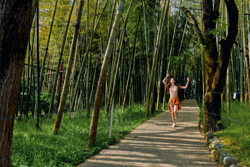 Bamboo forest path with woman walking barefoot in orange skirt, joyful arms raised on sunlit trail. Nature scene, serene walkway, green bamboo grove and peaceful outdoor lifestyle.