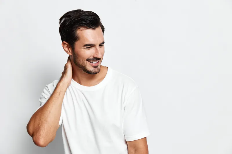 Smiling man wearing casual white t-shirt looking to side with a joyful expression while standing against a plain solid light gray background. People lifestyle concept.