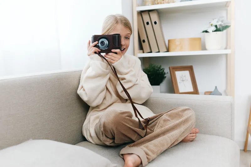 Young child sits on a light sofa holding a compact camera to their eye, ready to capture a moment. Cozy sweater, relaxed pose, warm natural light, soft home setting.
