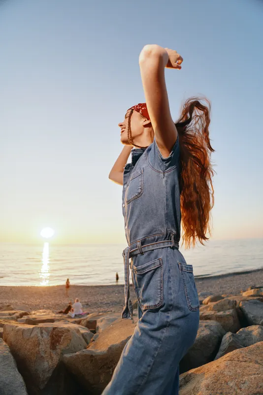 lifestyle woman in modern boho-western denim street style stretching by the beach at sunset with warm film color tones
