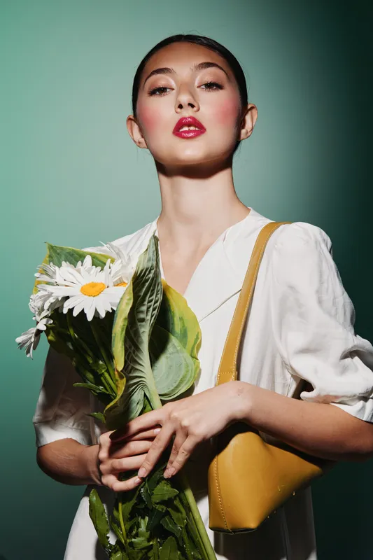 Young woman with bold red lipstick holding a large bouquet of daisies and greens, wearing a white blouse and yellow bag. Studio portrait on green background, focused on beauty and fashion.